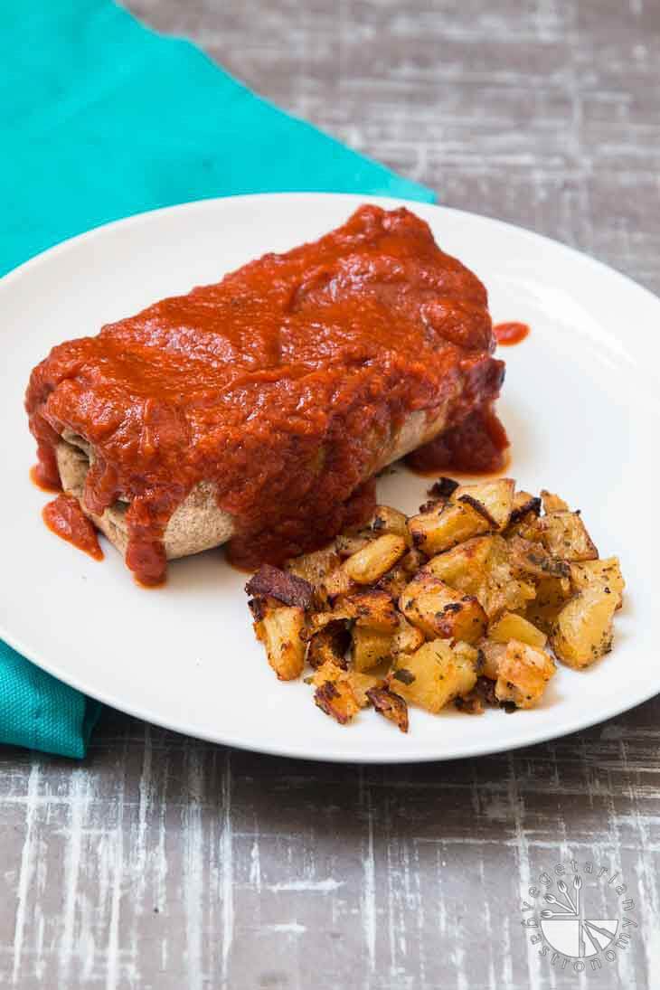 Side photograph of a breakfast burrito smoothered in red sauce, and served with a side of crispy potatoes. 