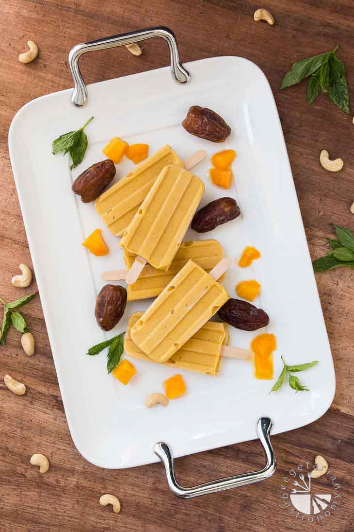 Overhead photograph of Vegan Mango Popsicles surrounded by dates, pieces of mangoes, cashews, and mint