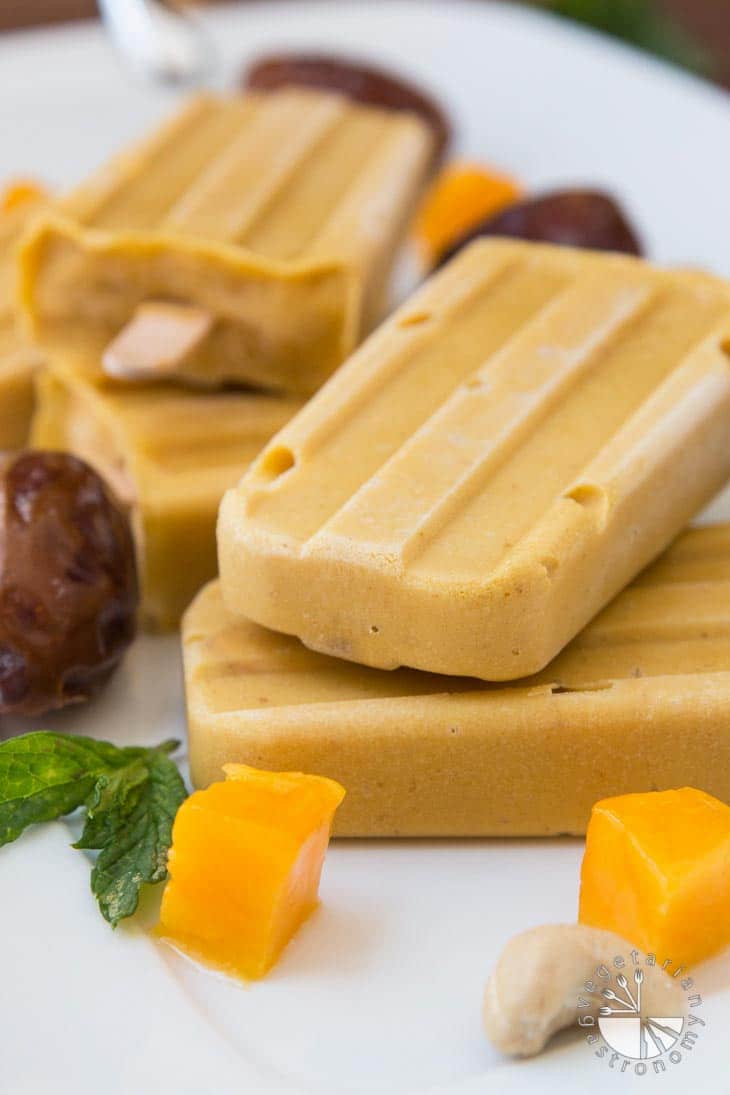 A closeup photograph of vegan mango popsicles on a white platter with pieces of dates, mint, and mango.