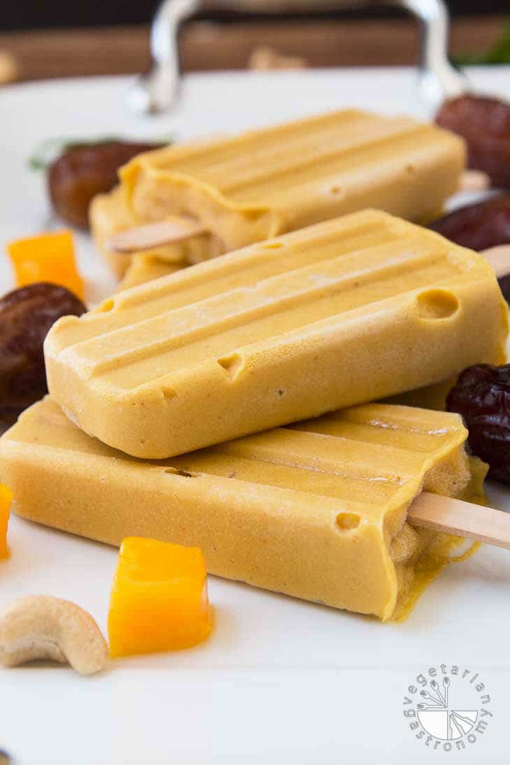 A side closeup photograph of vegan mango lassi popsicles surrounded by dates, cashews, and pieces of mango.