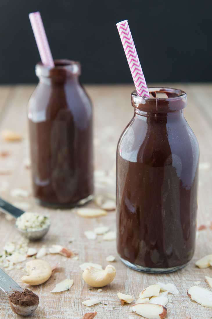 Front photograph of two milk bottles filled with vegan chocolate milk recipe. There are nuts and hemp seeds scattered in the background.
