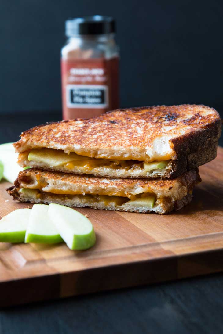 Pumpkin apple vegan grilled cheese cut in half and stalked on top of a brown cutting board with green apples off to the side. 