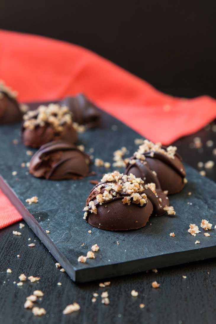 Vegan pumpkin spice dark chocolate truffles sprinkled with chopped pecans. The truffles are sitting on a black plate with a red napkin in the background.