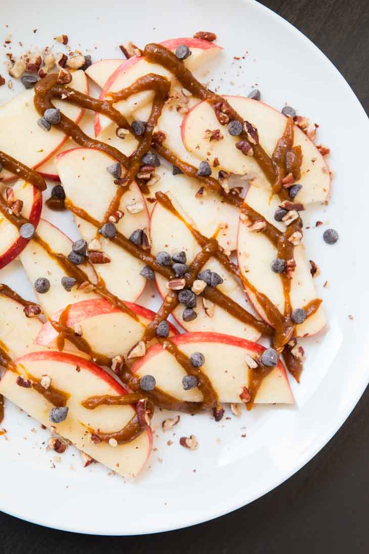 Overhead photograph of sliced apple nachos topped with pumpkin butter, chocolate, and pecans. They are sitting on a white round plate.