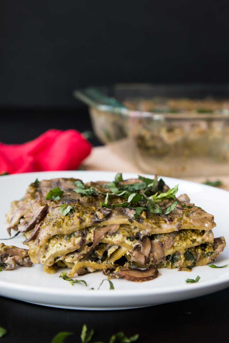 Ninth best vegan meal on VG in 2017. Side photograph of creamy spinach pesto lasagna with mushroom sauce sitting on a round white plate. There's a casserole dish and red napkin in the background. 