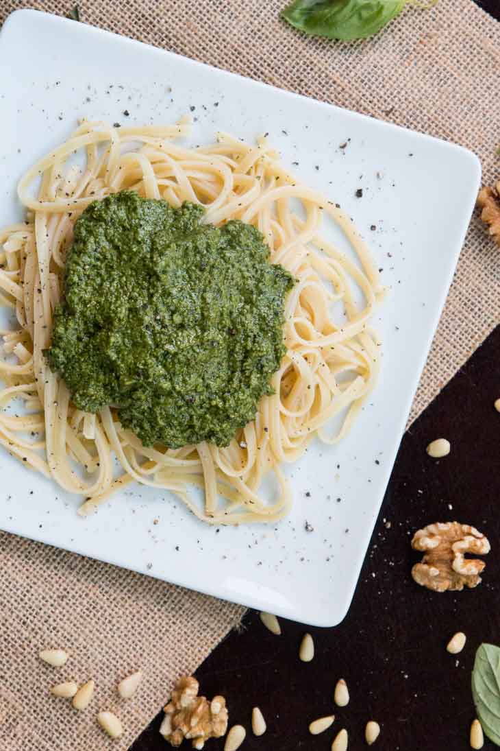 Fifth best vegan meal on VG in 2017. An overhead photograph of linguine pasta topped with vegan pesto, all plated on a square white plate. The table is garnished with nuts and basil. 