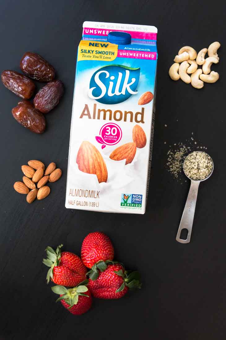 An overhead photograph showing the ingredients of the strawberry date milk recipe. There are dates, almonds, cashews, hemp seeds, strawberries, and silk almondmilk container.