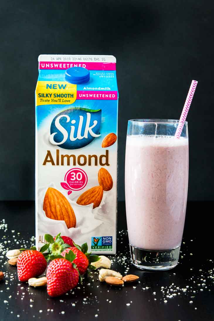 A photograph of a tall glass filled with nutty strawberry date milk and a pink straw. The glass is sitting next to a half gallon of silk almondmilk container. THere are strawberries, nuts, and hemp seeds scattered around.