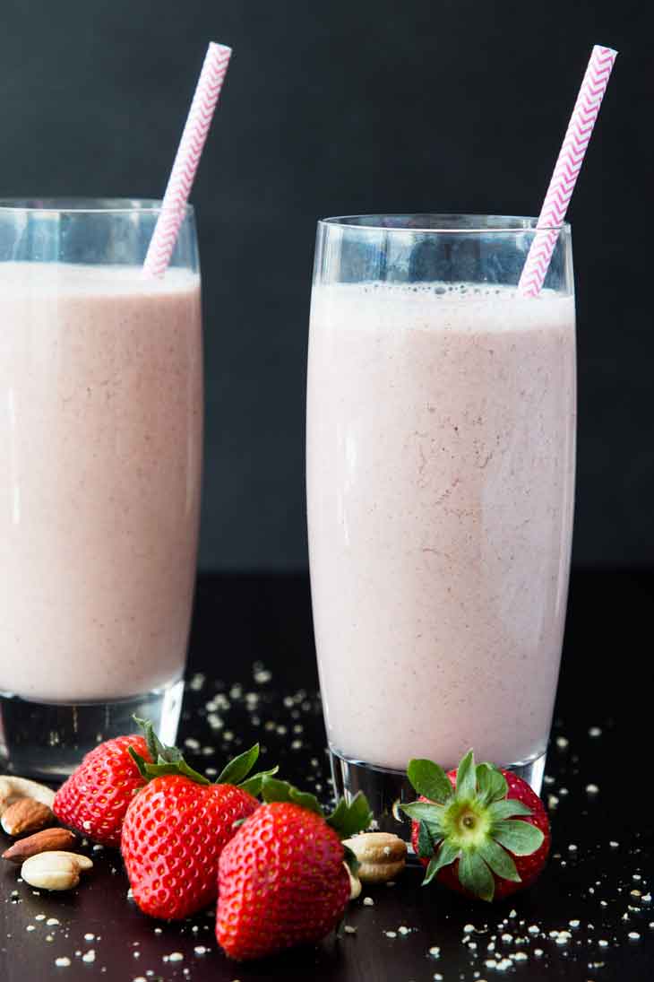 A photograph of two tall glasses of nutty strawberry date milk. There are strawberries, nuts, and hemp seeds scattered around.