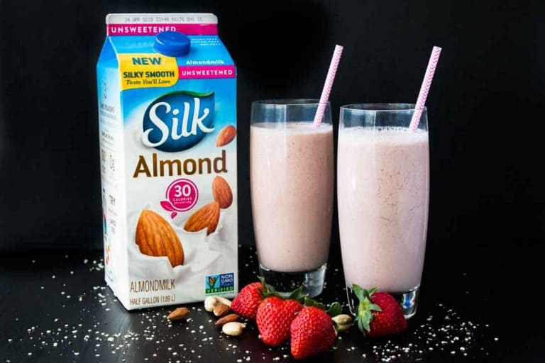 A side photograph of two tall glasses filled with strawberry date milk, containing pink straws. There's a container of silk almond milk, and strawberries, hemp seeds, and nuts scattered around.