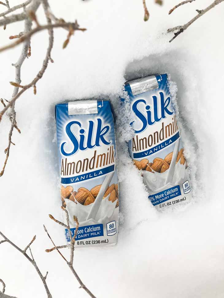 Two single serving silk almond milk containers sitting in the snow with branches on the side.