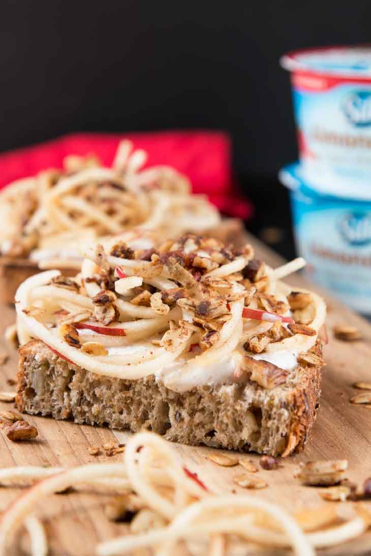 A close-up side photograph of vanilla yogurt cinnamon toast recipe on a cutting board with spiralized apples and granola. A red napkin and stalked almondmilk yogurt cups are in the background.