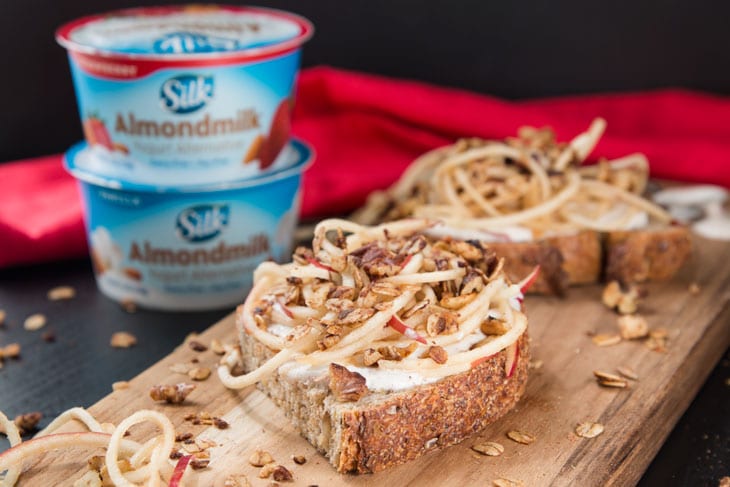 A side photograph of vanilla yogurt cinnamon toast recipe on a cutting board with spiralized apples and granola sprinkled on and around. There are two stacked single-serving SILK almondmilk yogurt cups in the background.