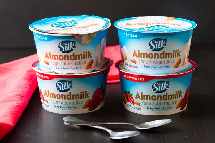 Photograph of 4 different single-serving SILK almond milk yogurt alternative cups. There are two spoons in front and a red napkin in the background.