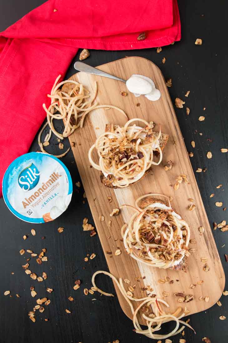 Overhead photograph of 2 cinnamon toast recipes served on a cutting board. There's a spoon with non-dairy yogurt, some granola sprinkled, almondmilk yogurt cup, and a red napkin off to the side.