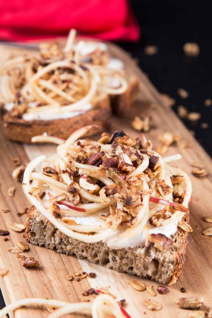 A 45 degree photograph of vanilla yogurt cinnamon toast recipe topped with spiralized apples and granola, served on a cutting board. There's a red napkin in the background.