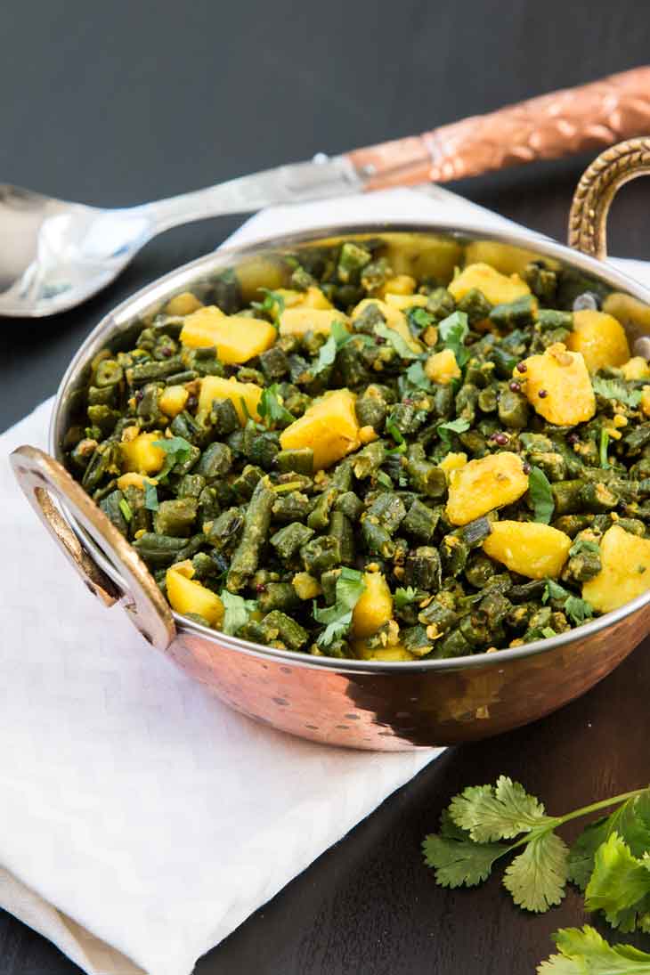 A side photograph of indian green bean curry with potatoes served in a copper bowl, alongside a white napkin and fresh cilantro. 