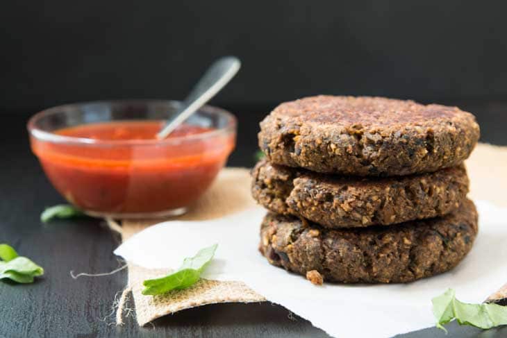 Side photograph of 3 vegan burger recipe patties stacked. There's a glass bowl of pizza sauce for serving on the side. 