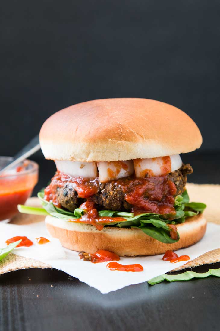 Perspective photograph of a vegan burger recipe with protein, lentils, onions, pizza sauce dripping off the sides, and spinach!