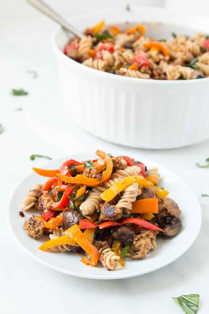 A white plate of vegan pasta salad with italian sausage, mushrooms, peppers, & cherry tomatoes. There's a serving plate in the background.