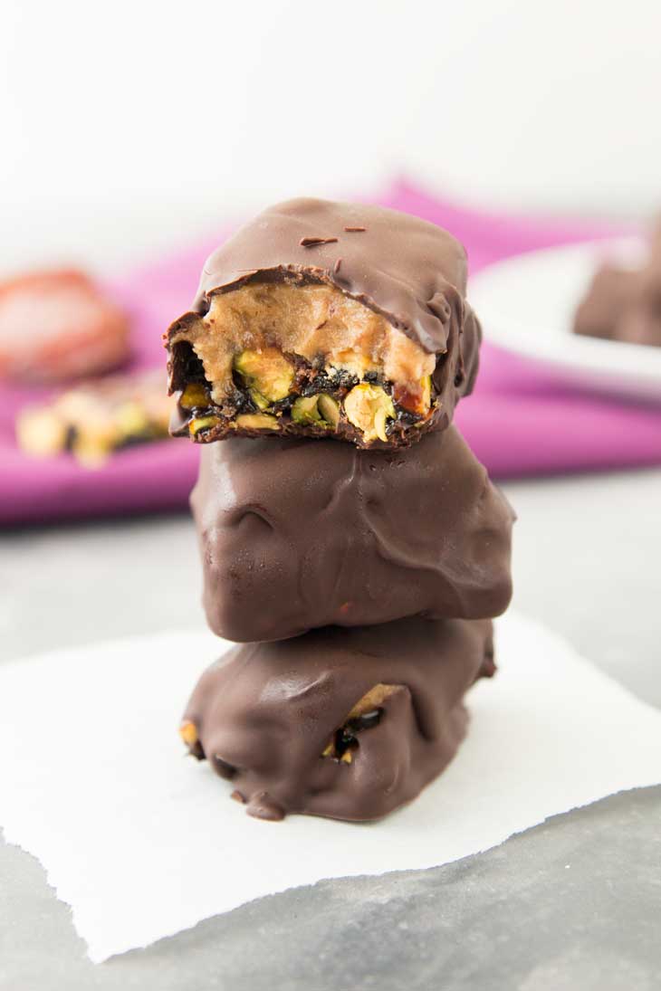 Three vegan chocolate bars stacked on parchment paper. The top bar is cut in half to show the date caramel and pistachios.