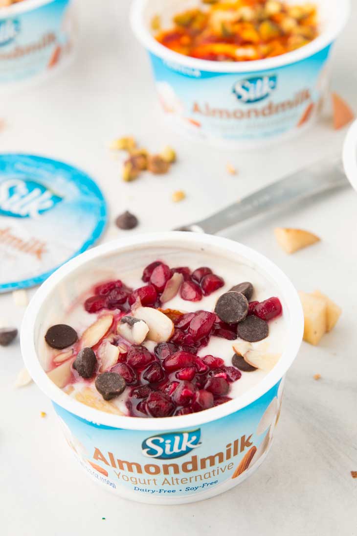 A side photograph showing a healthy yogurt cup with pomegranates, almonds, and dark chocolate. 