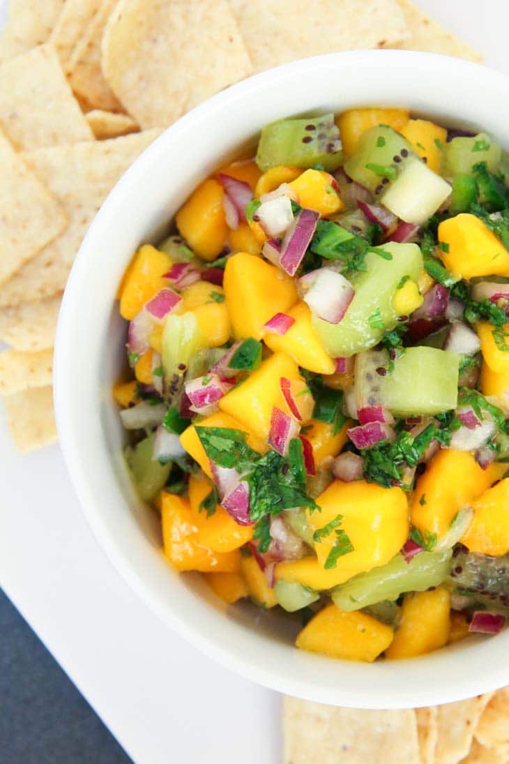 A close up of a mango fruit salsa in a white bowl with chips at the side