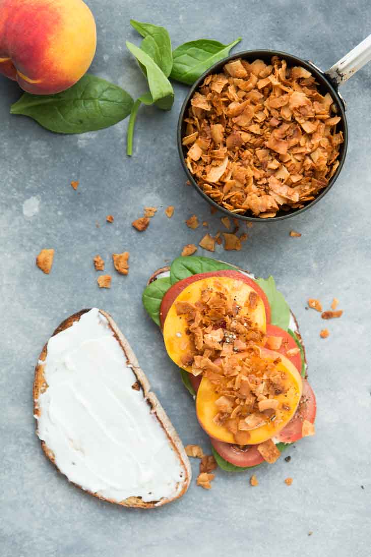 Overhead photograph of an open faced vegan BLT sandwich with peaches and coconut bacon. There's peaches, coconut bacon, and spinach leaves as garnish.