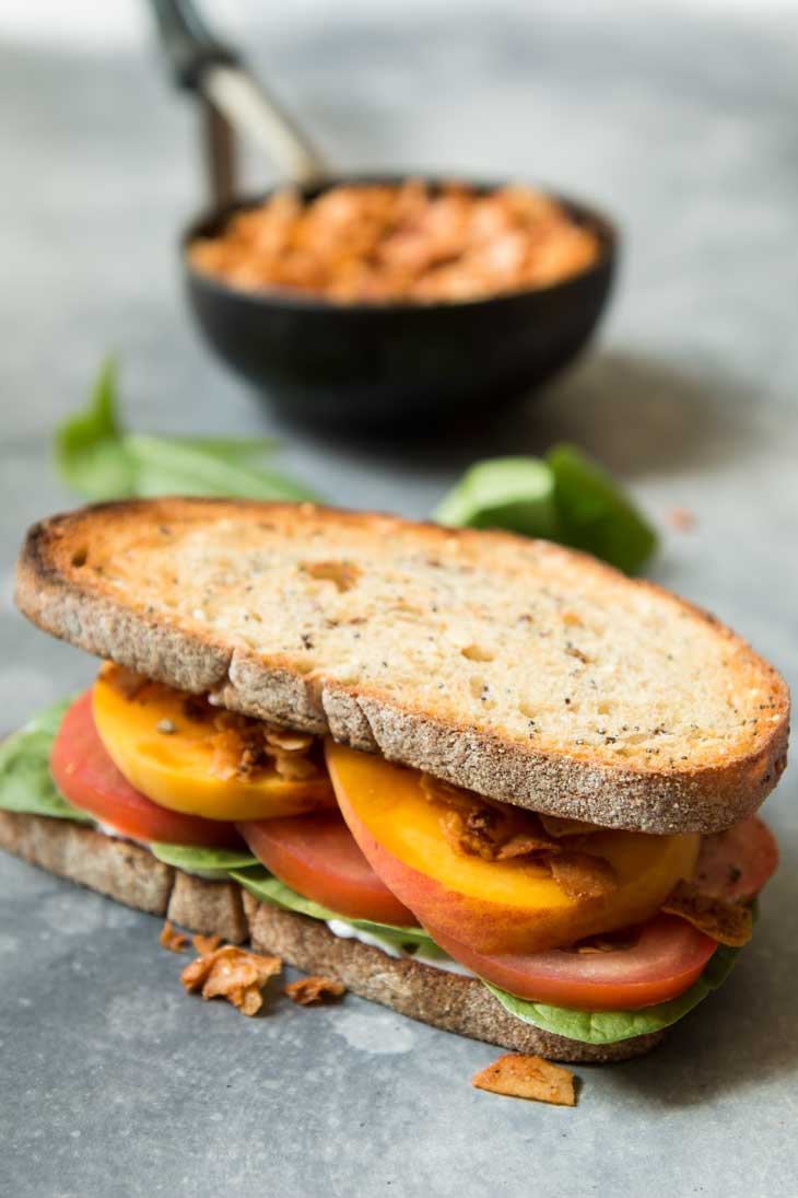 Side photoraph of vegan BLT sandwich with spinach, peaches, and coconut bacon. There's coconut bacon in the background.