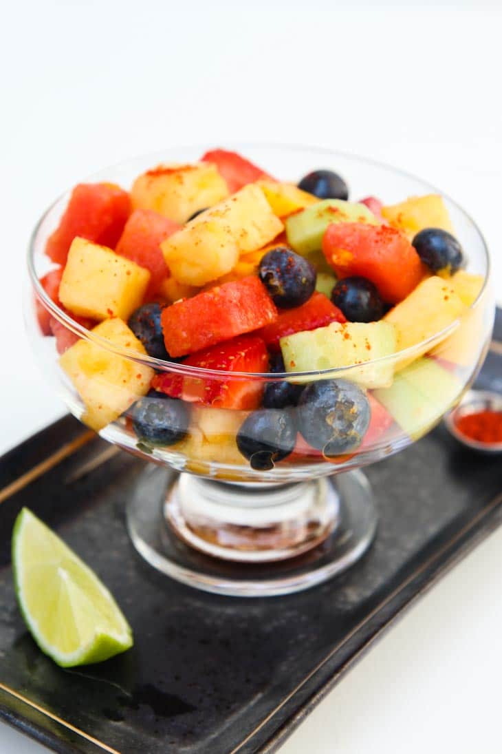 A large glass of homemade fruit salad on a tray with a lime wedge at the side