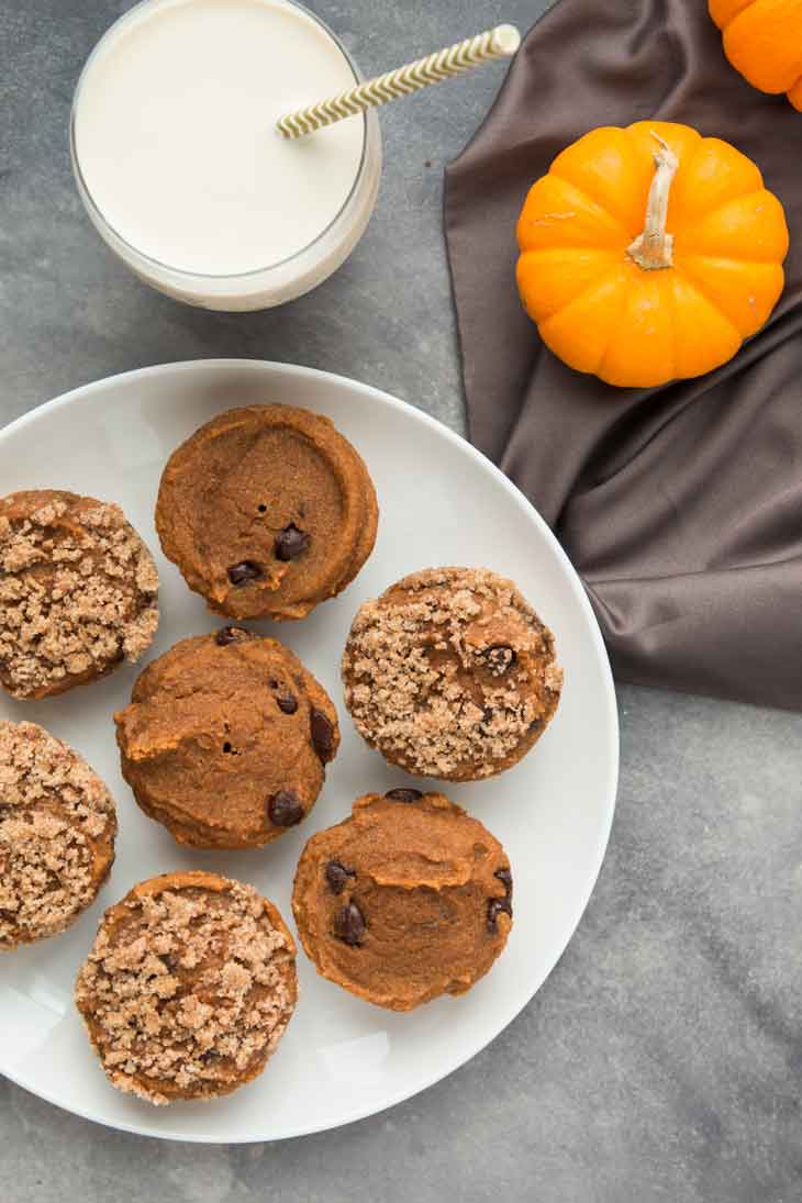 Overhead photograph of vegan pumpkin muffins on a white plate. There's a glass of vegan milk and some mini pumpkins on the side.