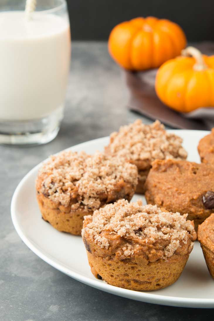 A side close-up of vegan pumpkin muffins on a white plate. There's a glass of milk and mini pumpkins in the background.