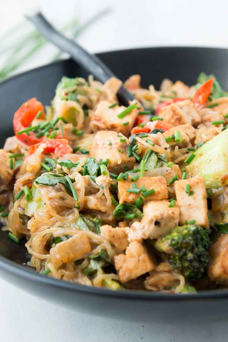 Side close-up of a healthy stir fry noodle with tofu and veggies in a black bowl with a spoon and chives in the background.