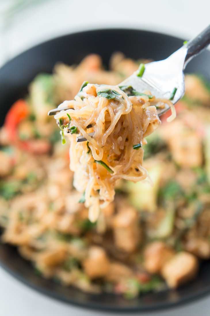 A close up photograph of healthy stir fry noodles being scooped up by a fork with the bowl in the background.