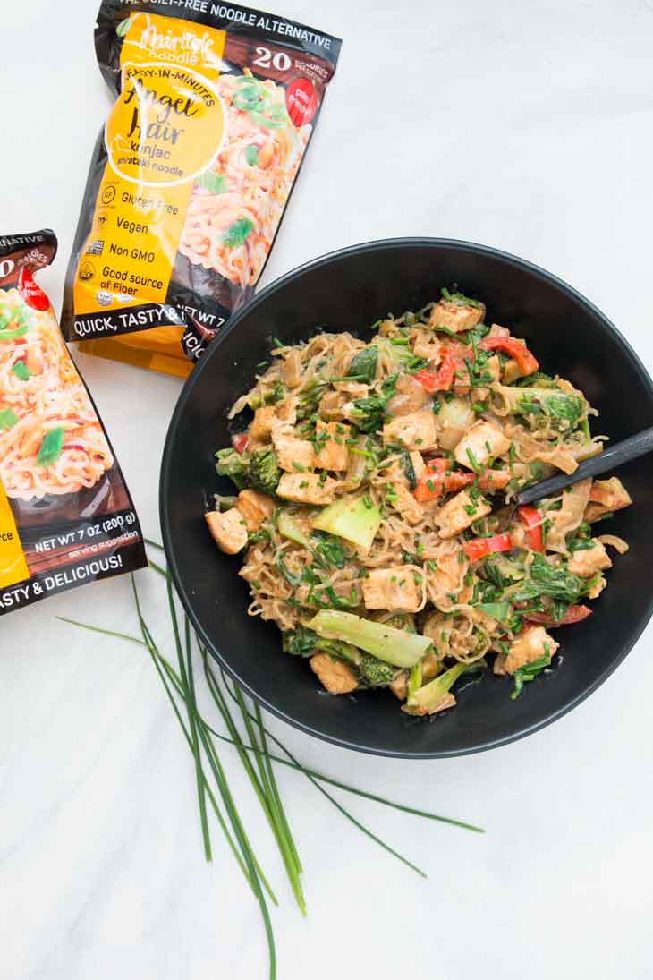 Overhead photograph of healthy stir fry noodles with tofu and veggies. There are packages of low-carb noodles and chives on the side.