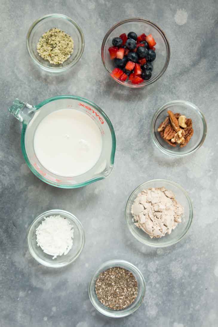 An overhead photograph of all the ingredients needed to make a vegan keto superfood breakfast bowl, including non-dairy milk, seeds, protein powder, mixed berries, and nuts.