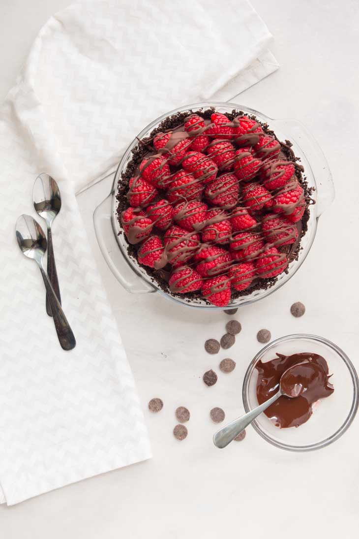 An overhead photograph of vegan chocolate pie topped with raspberries. There's a bowl of melted chocolate, chocolate chips, and spoons off to the side.