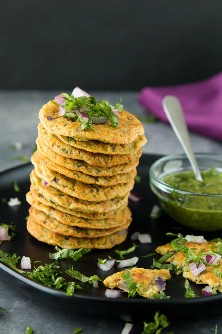 A stack of mini vegetable pancakes on a black plate, with a green pesto dipping sauce, onions, and fresh cilantro!