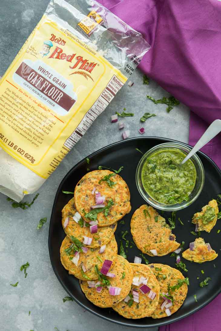 Overhead photograph of savory mini vegetable pancakes with a bag of Bob's Red Mill oat flour on the side. There's a green sauce for dipping and scattered herbs.