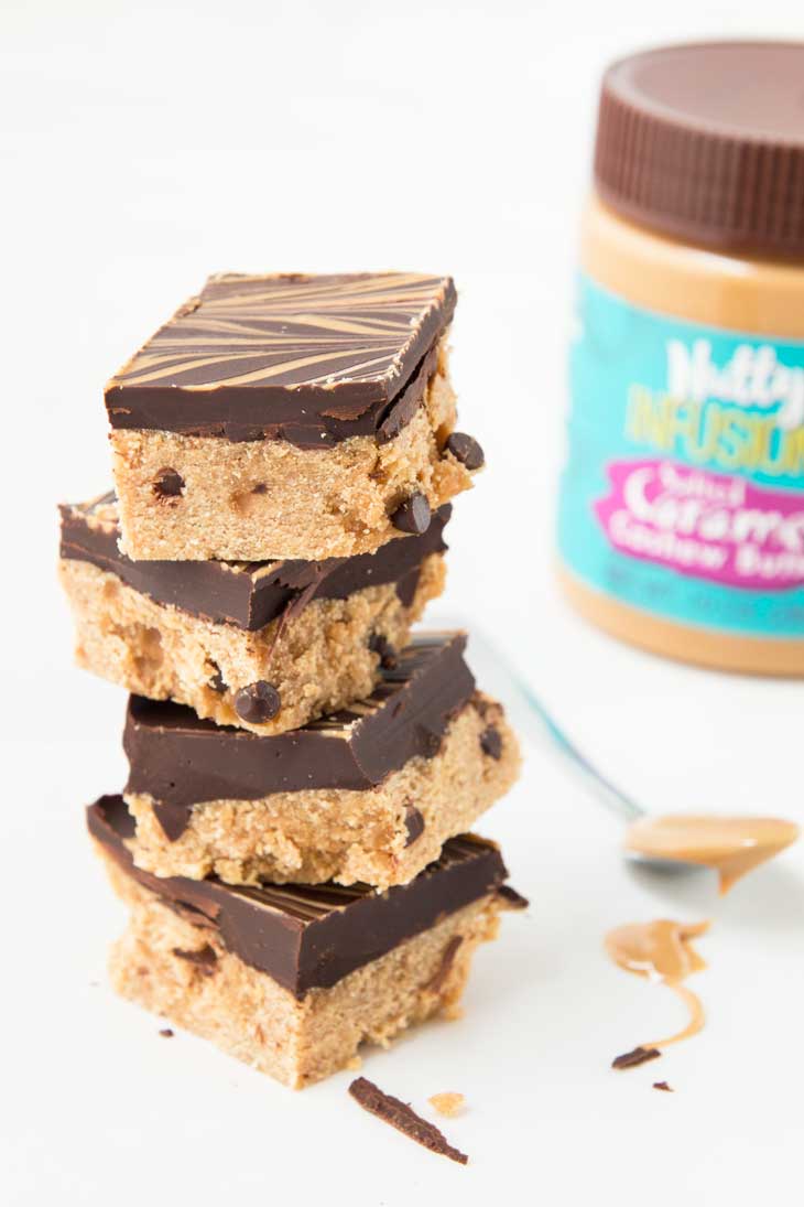 A stack of salted caramel bars, with a spoon full of cashew butter, and a container in the background. 