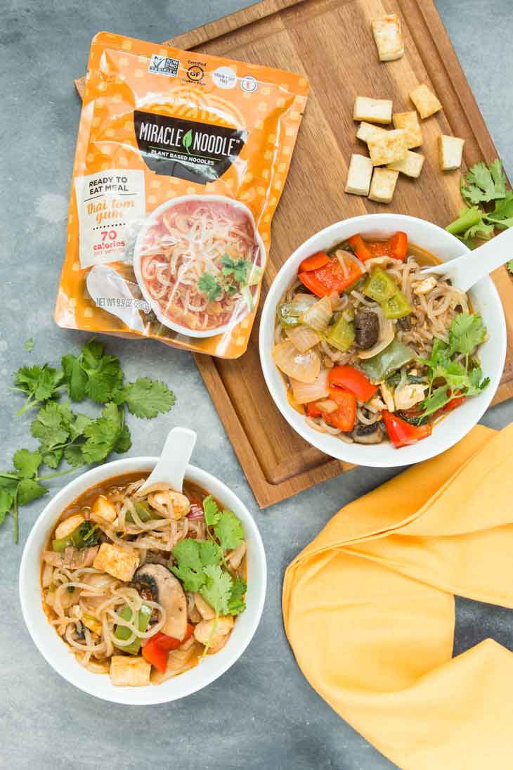 Overhead photograph of two white bowls with tom yum thai vegan noodle soup containing vegetables and tofu. There's fresh cilantro and miracle noodle pack on the side.