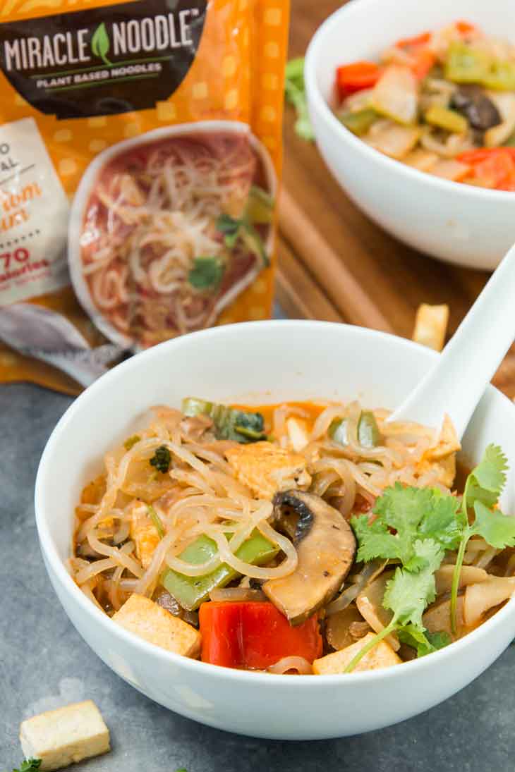 Side photograph of thai tom yum vegan noodle soup with vegetables and fried tofu. There's a package of miracle noodle in the background.