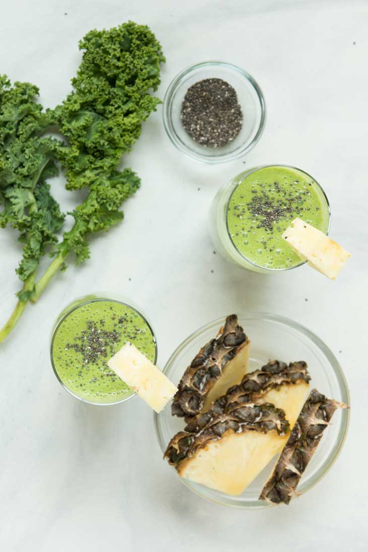 Overhead shot of fresh kale leaves, two pineapple kale smoothie, fresh pineapple, and a bowl of chia seeds.