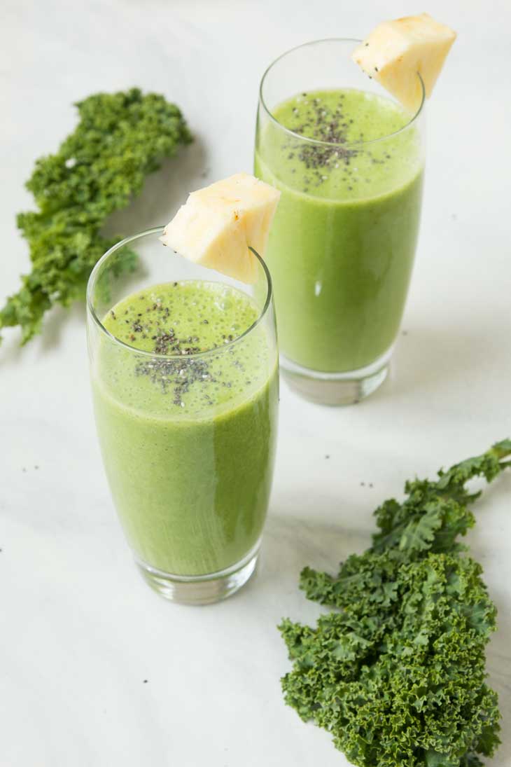 Side photograph of two glasses filled with pineapple kale smoothie, and fresh kale leaves on the side.